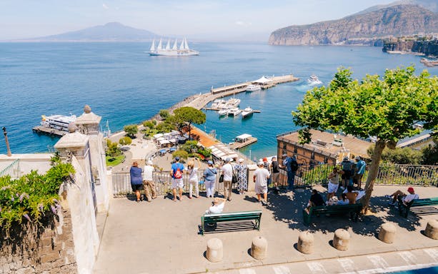 Tourists overlooking Marina Piccola in Sorrento with boats and Mount Vesuvius in the background.