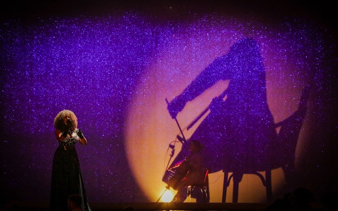 Performer singing with shadow of cellist at Paradis Latin festive show.