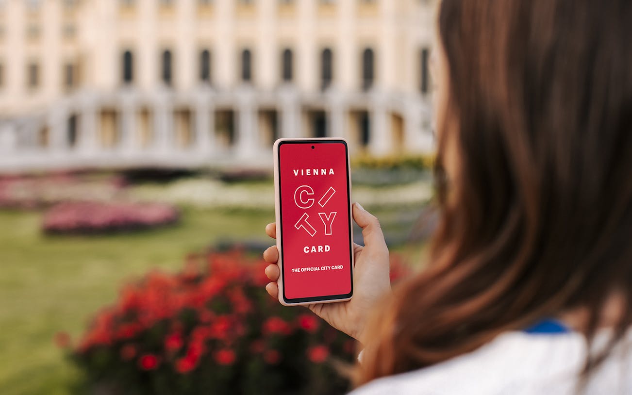 Person holding a phone displaying Vienna City Card with a blurred historic building in the background.
