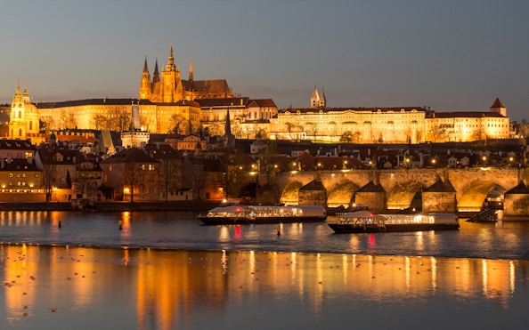 Open-top glass boat on Vltava River with Prague Castle illuminated at night.