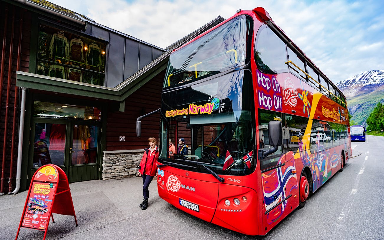 Red double-decker bus for Geiranger Hop-on Hop-off tour parked by a building in Norway.