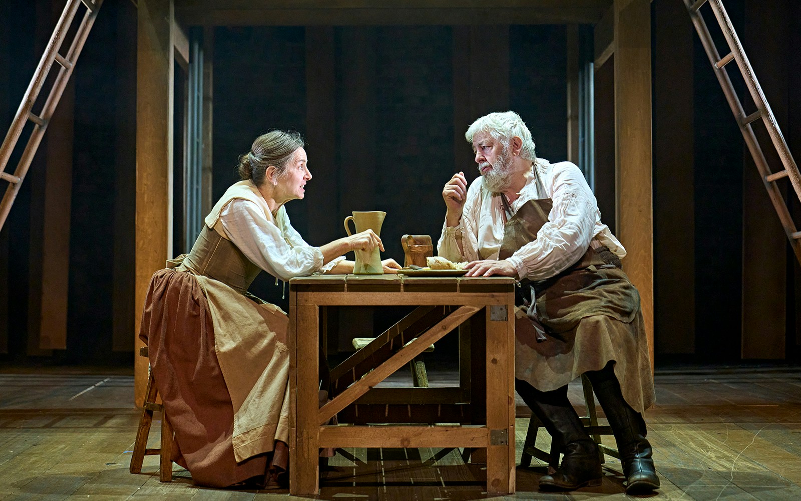 Two actors in period costumes perform a scene from "Hamnet" at a wooden table on stage.