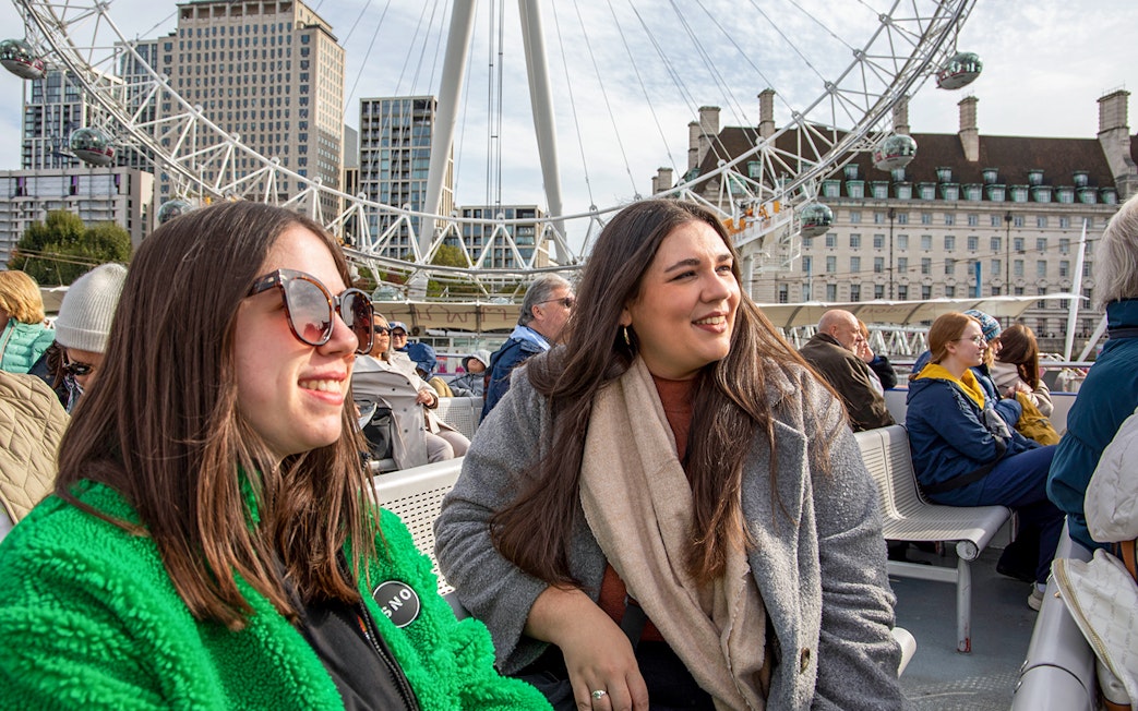 Women enjoying Thames River cruise with London Eye in background.