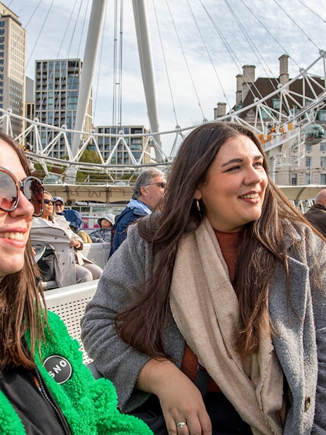 Women enjoying Thames River cruise with London Eye in background.