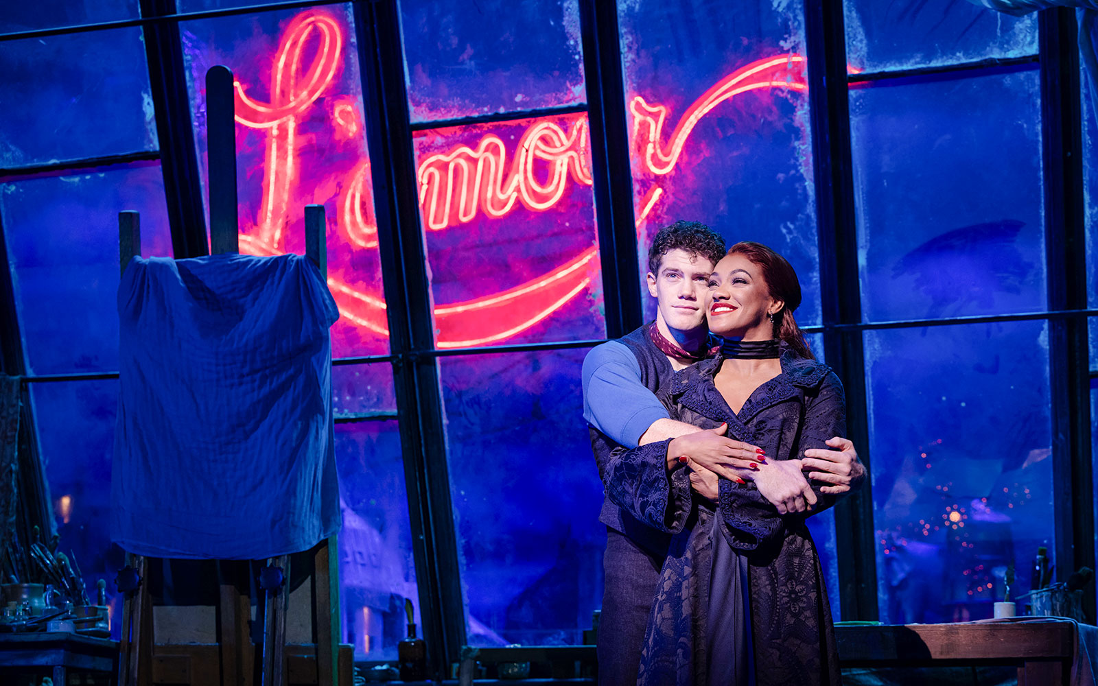 Couple embracing on stage during Moulin Rouge! The Musical in West End with neon "Amour" sign.