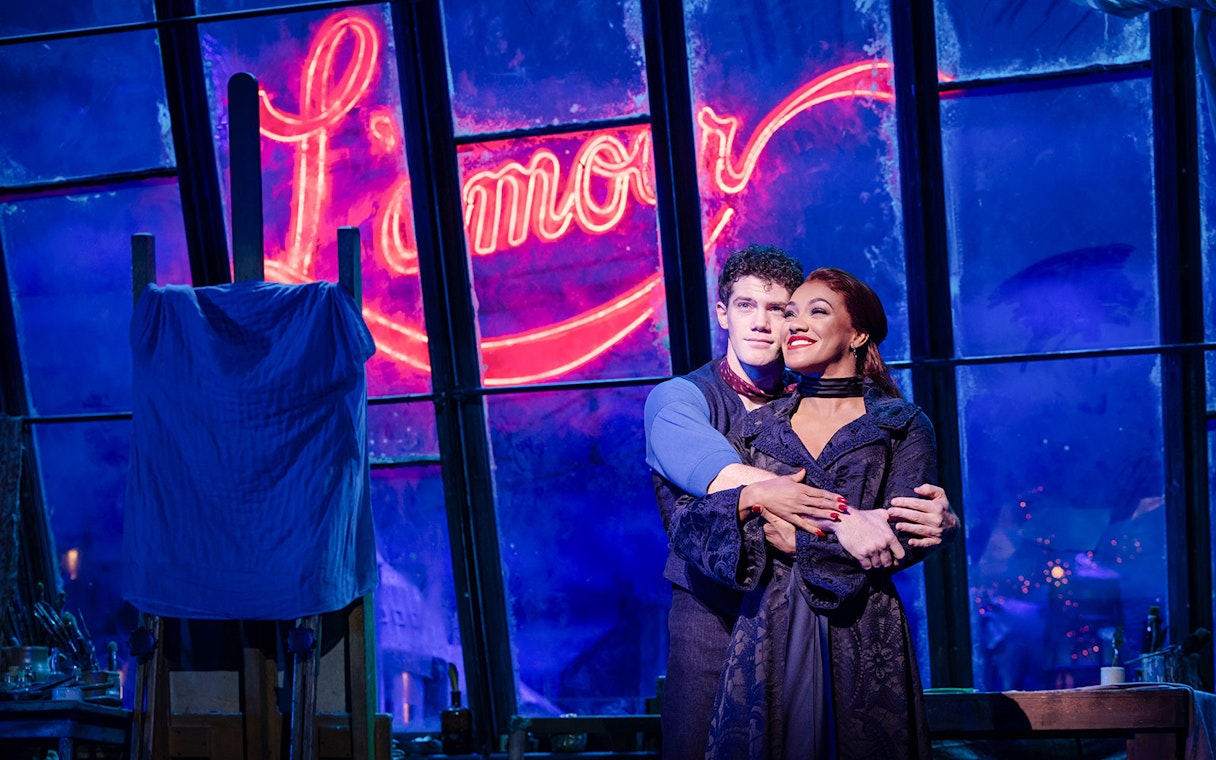 Couple embracing on stage during Moulin Rouge! The Musical in West End with neon "Amour" sign.