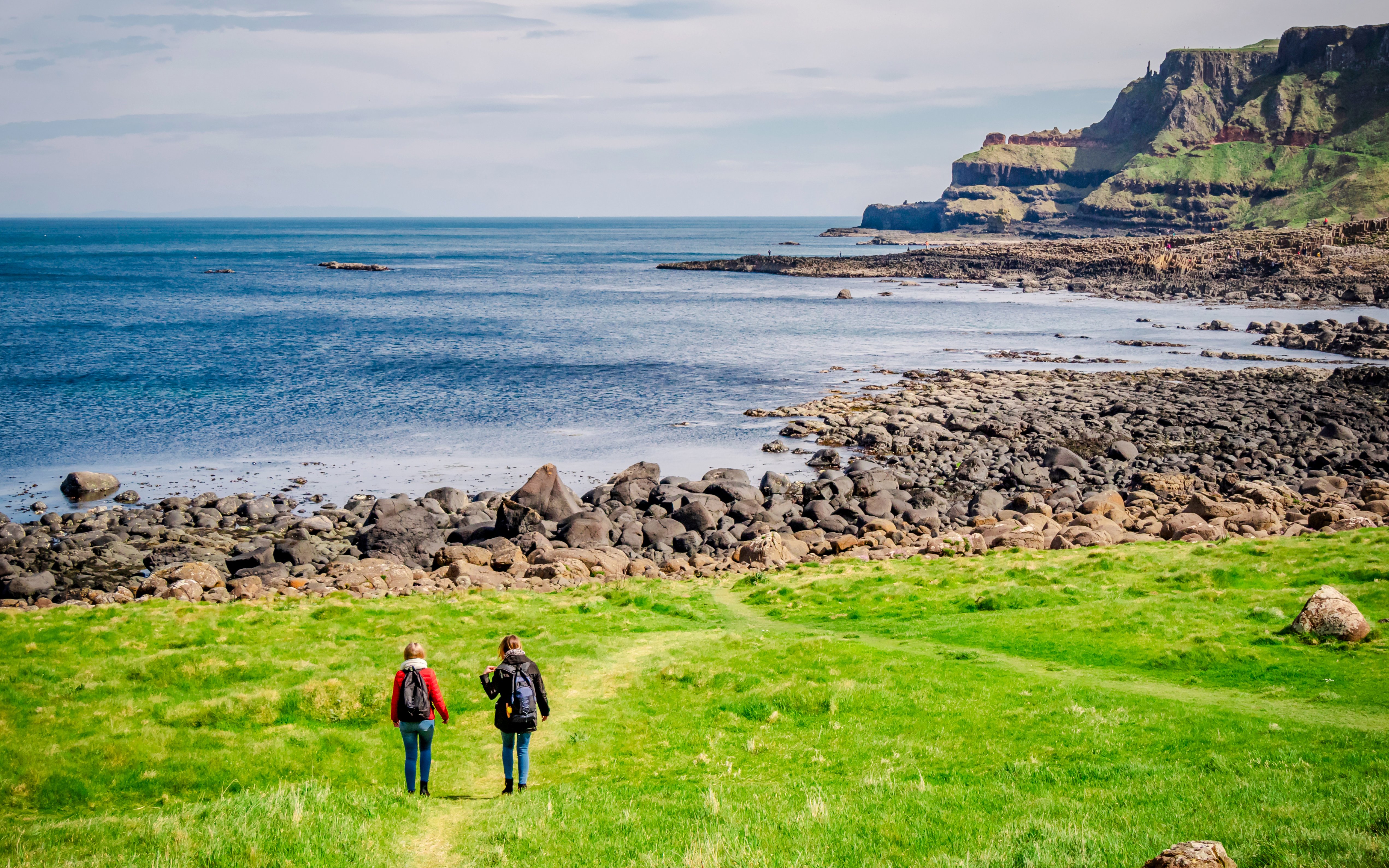 Two people walking on a grassy path towards the rocky coastline of Giant’s Causeway, Northern Ireland.