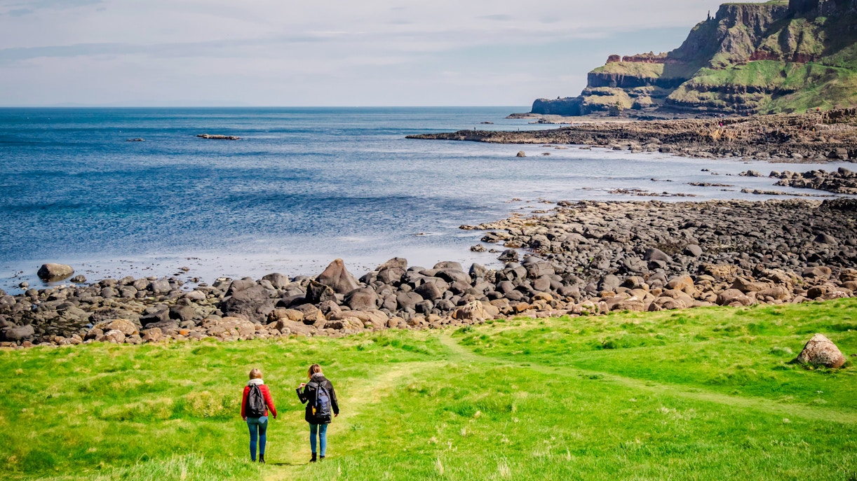 Two people walking on a grassy path towards the rocky coastline of Giant’s Causeway, Northern Ireland.