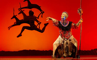 Lion King performer in vibrant costume on West End stage with antelope silhouette.