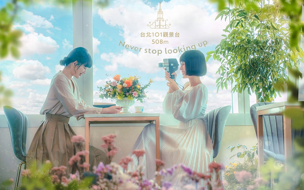 Two women enjoying a view from Taipei 101 observatory with flowers and a camera.