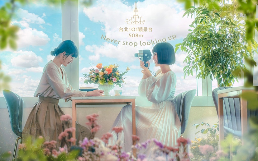 Two women enjoying a view from Taipei 101 observatory with flowers and a camera.