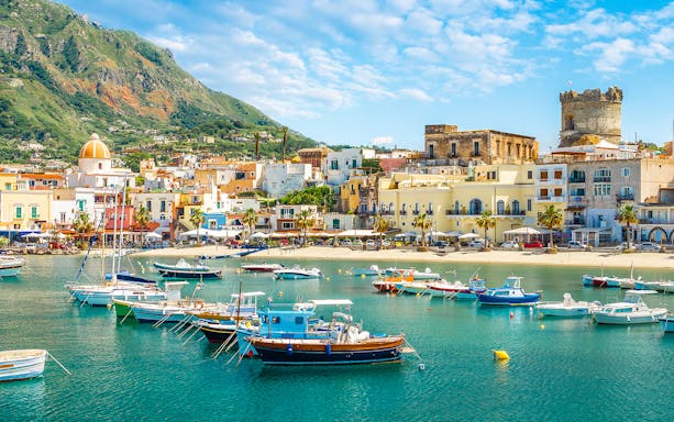 Boats in the harbor of Ischia with colorful buildings and mountains in the background.