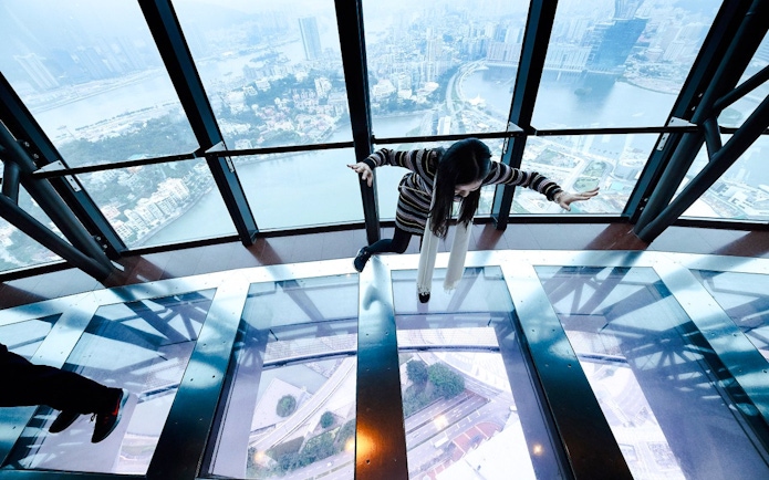 Tourist balancing on glass floor at Macau Tower Observation Deck with city view.