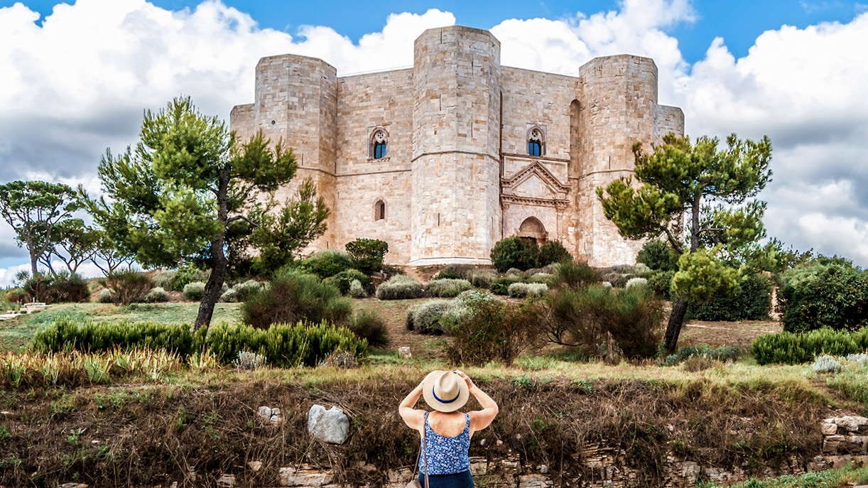 A women tourist with her traveller hat on, clicking a photo of the Castel from the outer area.