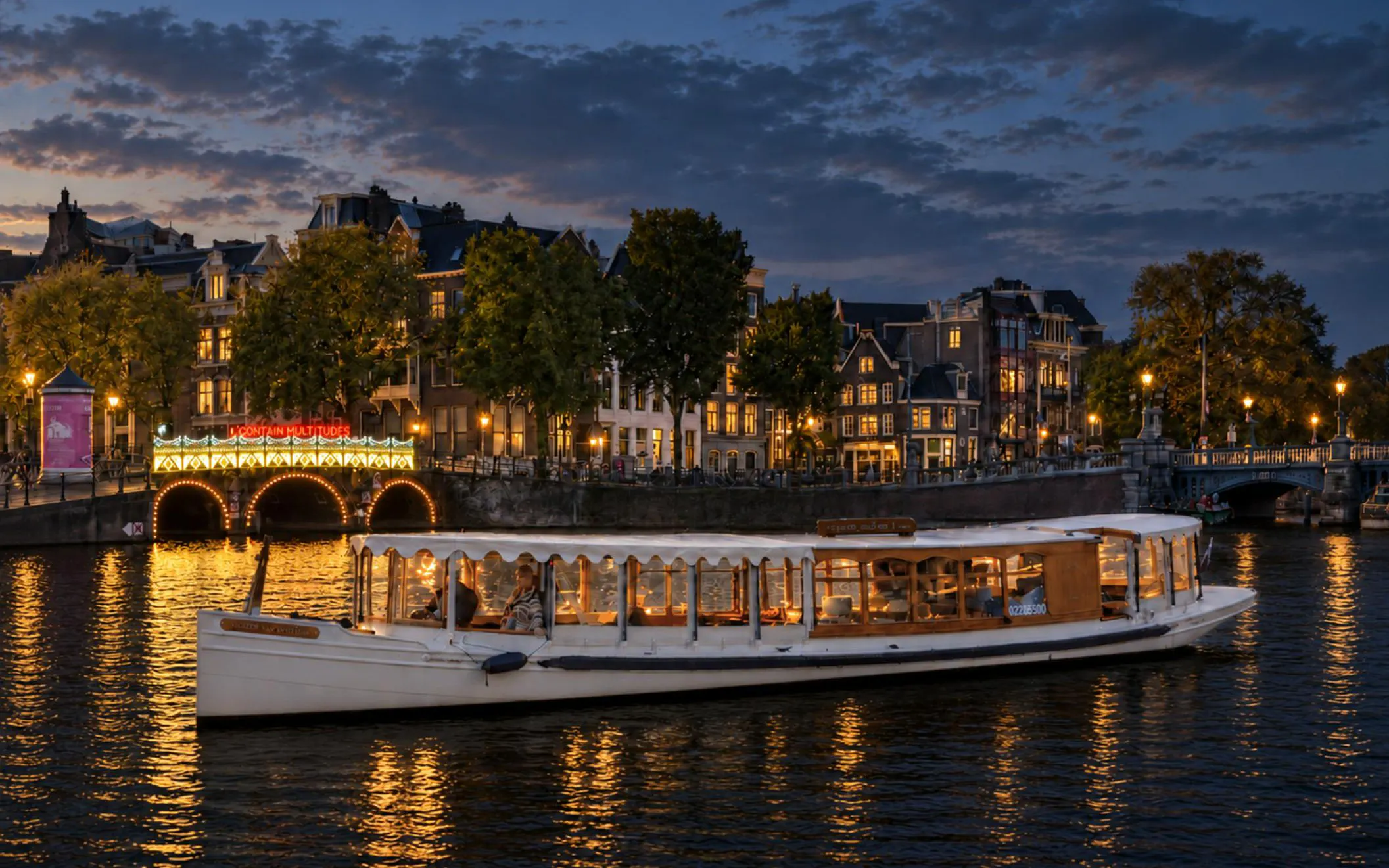 Canal boat cruising during Amsterdam Light Festival with illuminated bridge and historic buildings.