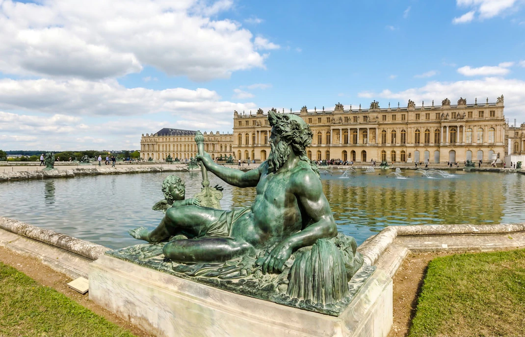 Statue in front of the Palace of Versailles with reflecting pool and building facade.