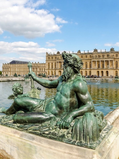 Statue in front of the Palace of Versailles with reflecting pool and building facade.