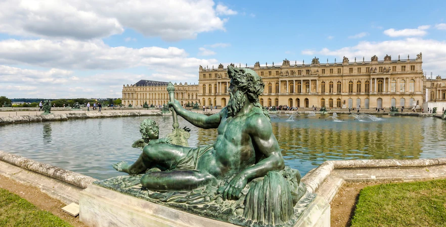 Statue in front of the Palace of Versailles with reflecting pool and building facade.