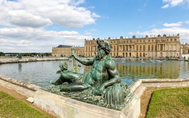 Statue in front of the Palace of Versailles with reflecting pool and building facade.