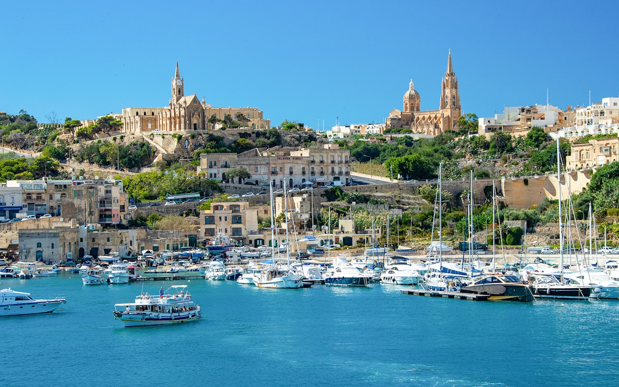 Harbor view of Gozo with boats and historic buildings on a hillside.