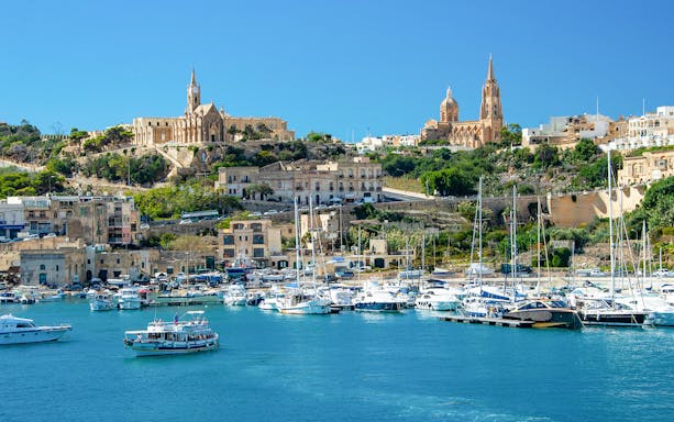 Harbor view of Gozo with boats and historic buildings on a hillside.
