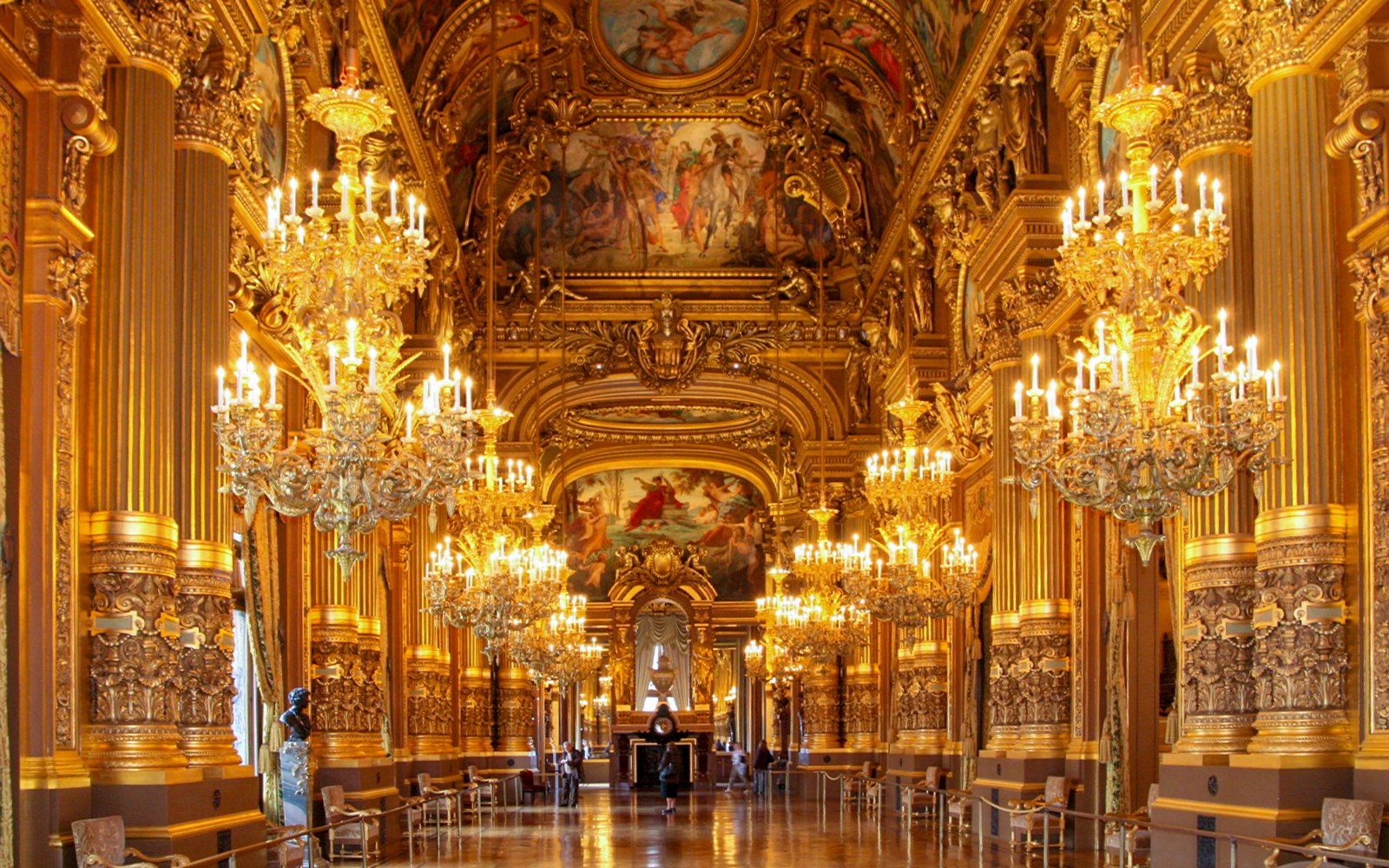 Visitors marveling at the ornate gold detailing and grand chandeliers in the Grand Foyer of the Opera Garnier in Paris, France