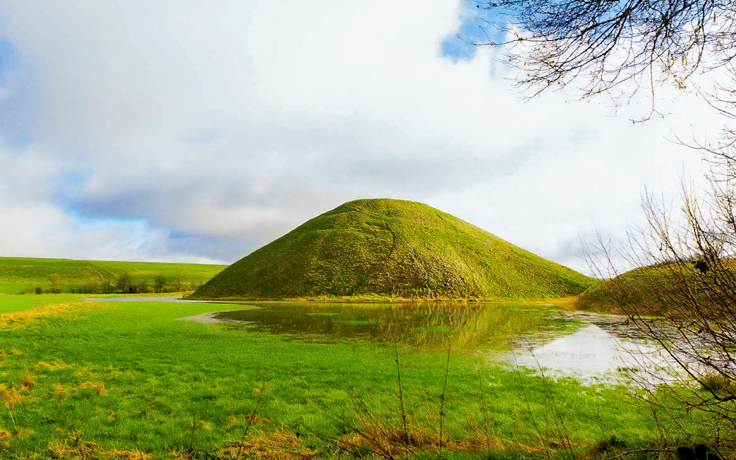 Silbury Hill in Avebury, England, surrounded by green fields and a reflective pond.