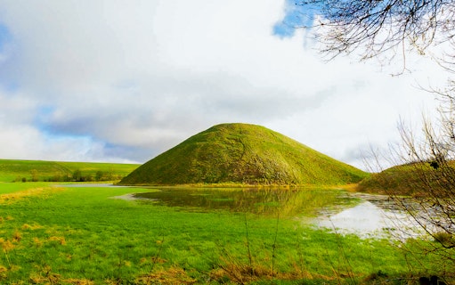 Silbury Hill in Avebury, England, surrounded by green fields and a reflective pond.
