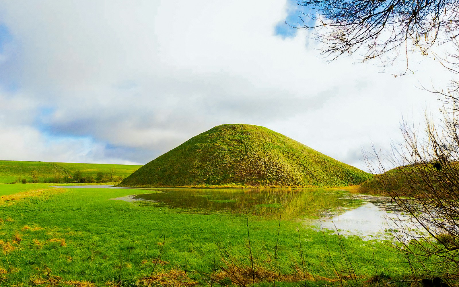 Silbury Hill in Avebury, England, surrounded by green fields and a reflective pond.