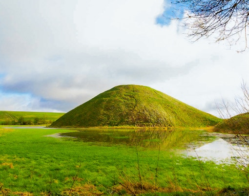 Silbury Hill in Avebury, England, surrounded by green fields and a reflective pond.
