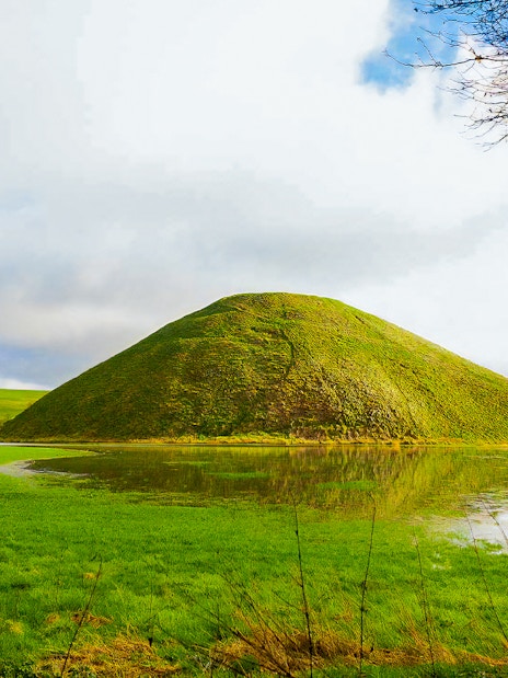 Silbury Hill in Avebury, England, surrounded by green fields and a reflective pond.