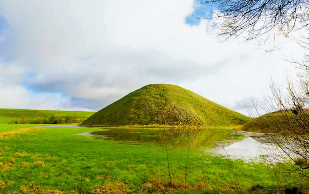 Silbury Hill in Avebury, England, surrounded by green fields and a reflective pond.