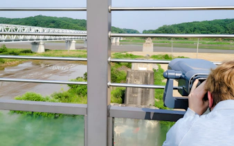 Person using binoculars to view the DMZ landscape with a bridge in the background.