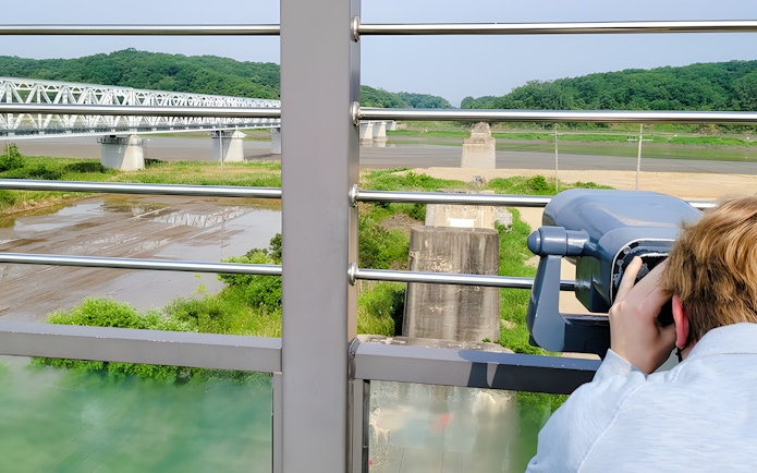 Person using binoculars to view the DMZ landscape with a bridge in the background.
