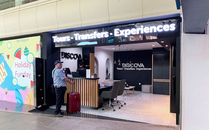 Man with suitcase at Discova Tours counter, Bali Airport.