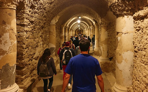 Tour group exploring ancient Roman catacombs with stone columns and arched ceilings.