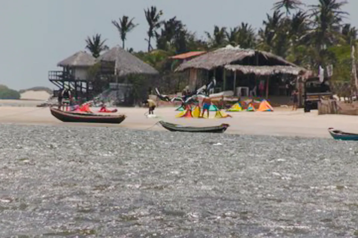 Kitesurfers and boats on the beach at Canto do Atins, Sao Paulo, with palm trees and huts in the background.
