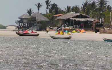 Kitesurfers and boats on the beach at Canto do Atins, Sao Paulo, with palm trees and huts in the background.