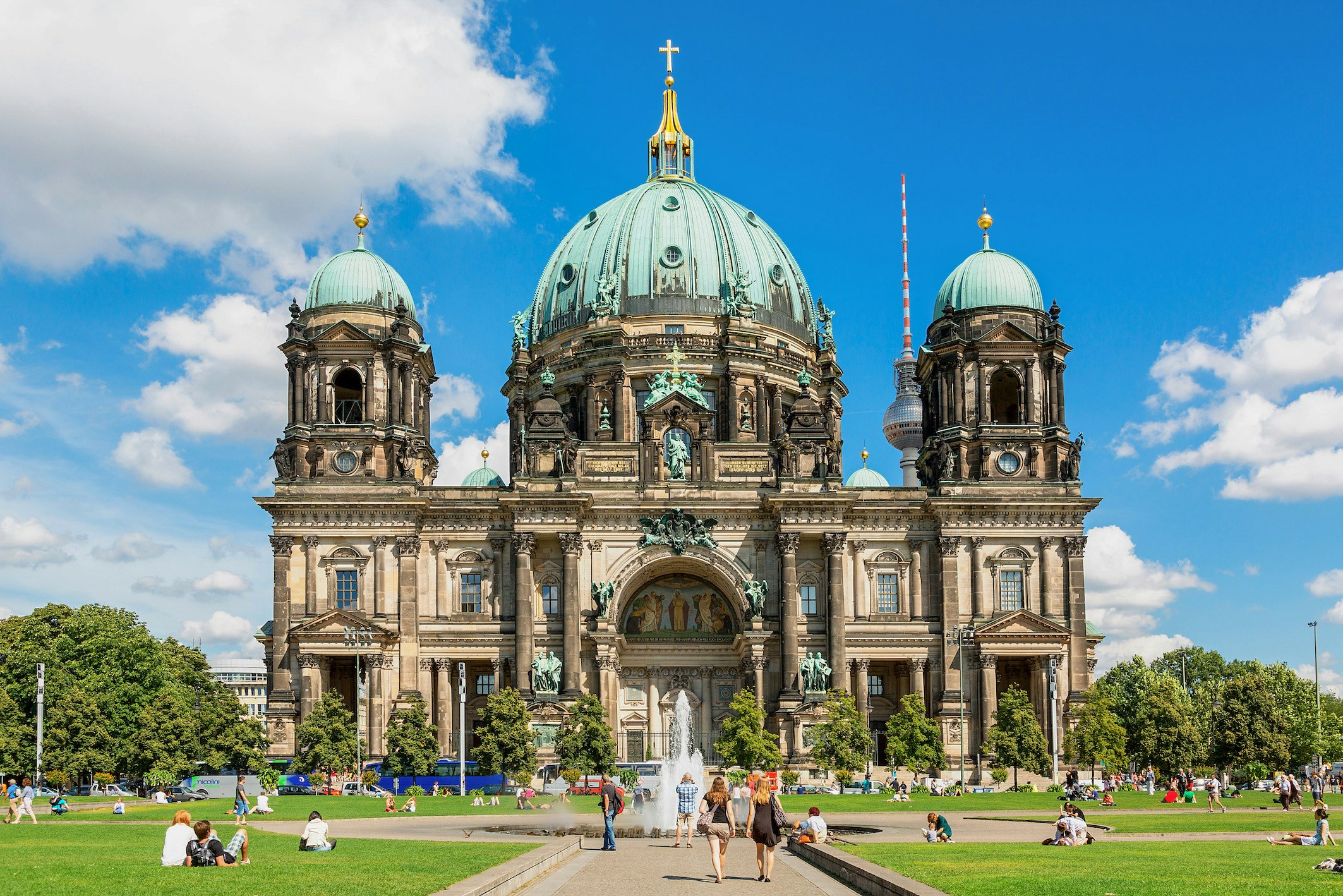 Berlin Cathedral exterior with TV Tower in background, people relaxing on lawn.
