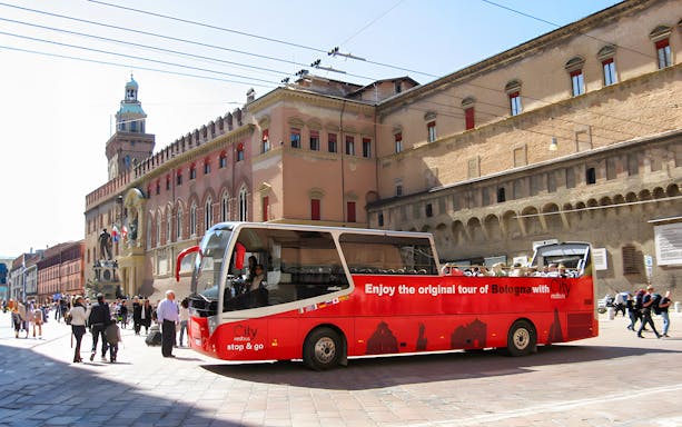 Red city tour bus in Bologna near historic buildings.