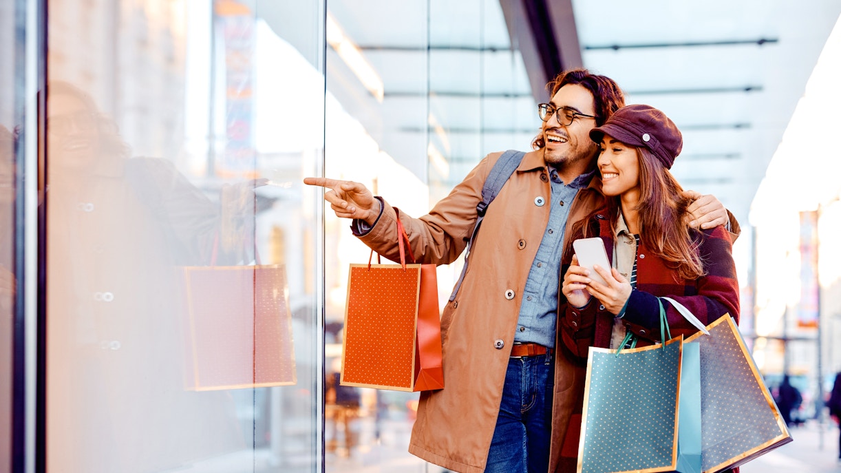 Couple shopping at La Vallee Village, Paris, holding bags and pointing at a store window.