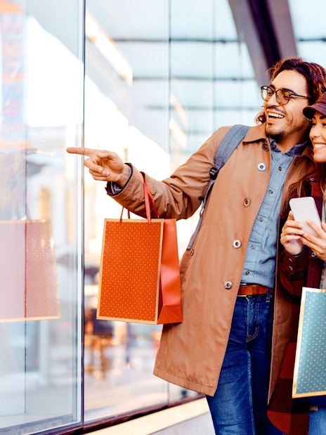 Couple shopping at La Vallee Village, Paris, holding bags and pointing at a store window.