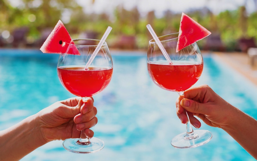 Two hands holding glasses of red drinks with watermelon slices by a pool.
