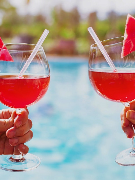 Two hands holding glasses of red drinks with watermelon slices by a pool.