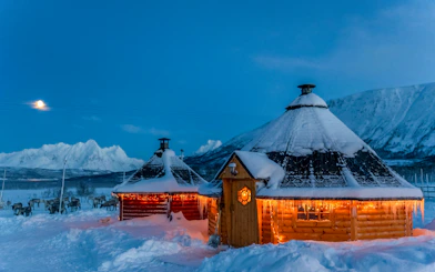 Snow-covered Sami huts with reindeer in Tromso under moonlit sky.