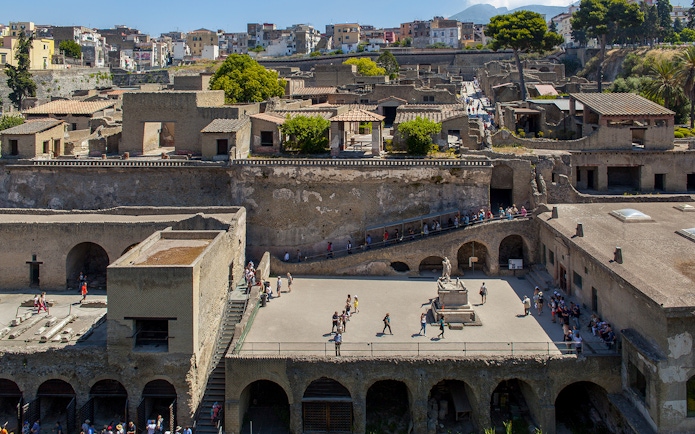 People walking through the ancient ruins of Herculaneum, Sorrento, with historic buildings in the background.