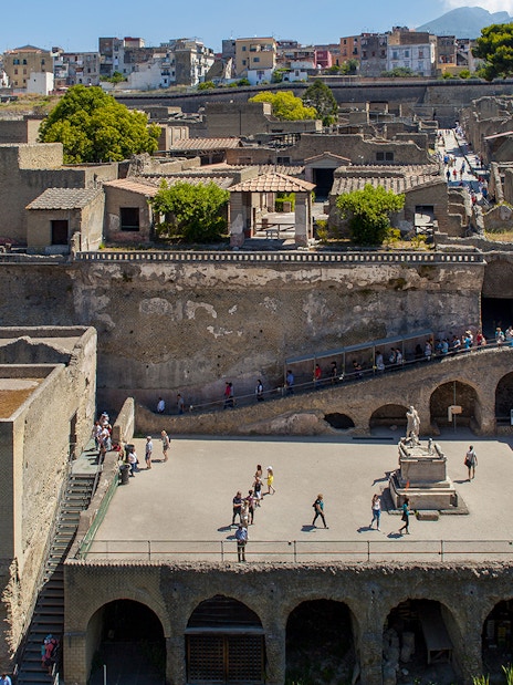 People walking through the ancient ruins of Herculaneum, Sorrento, with historic buildings in the background.