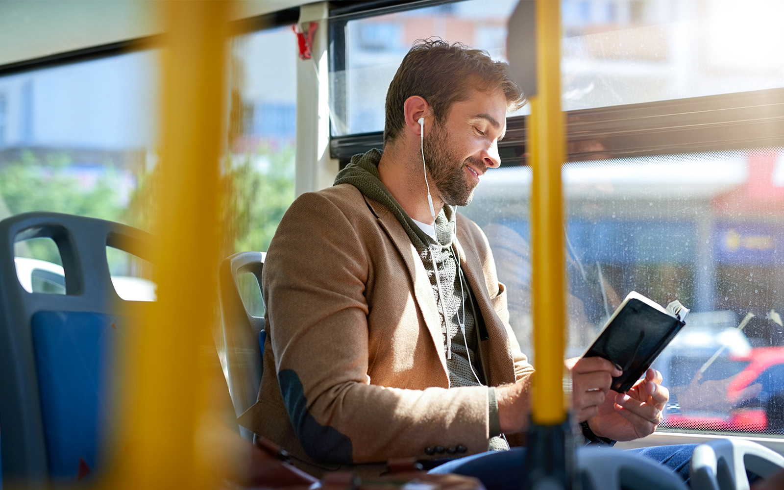 Man reading a book on a bus while listening to earphones.