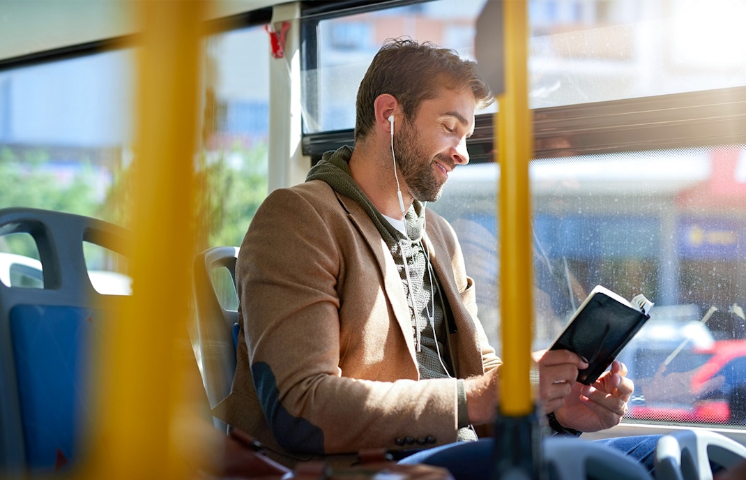 Man reading a book on a bus while listening to earphones.