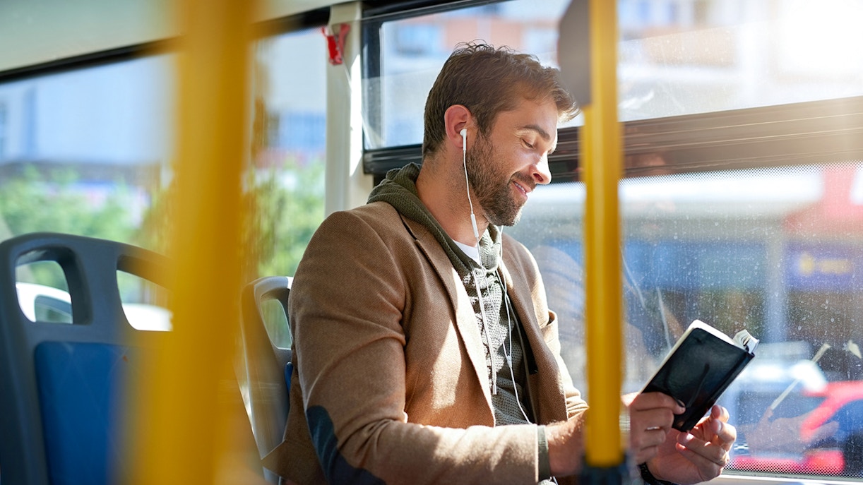 Man reading a book on a bus while listening to earphones.